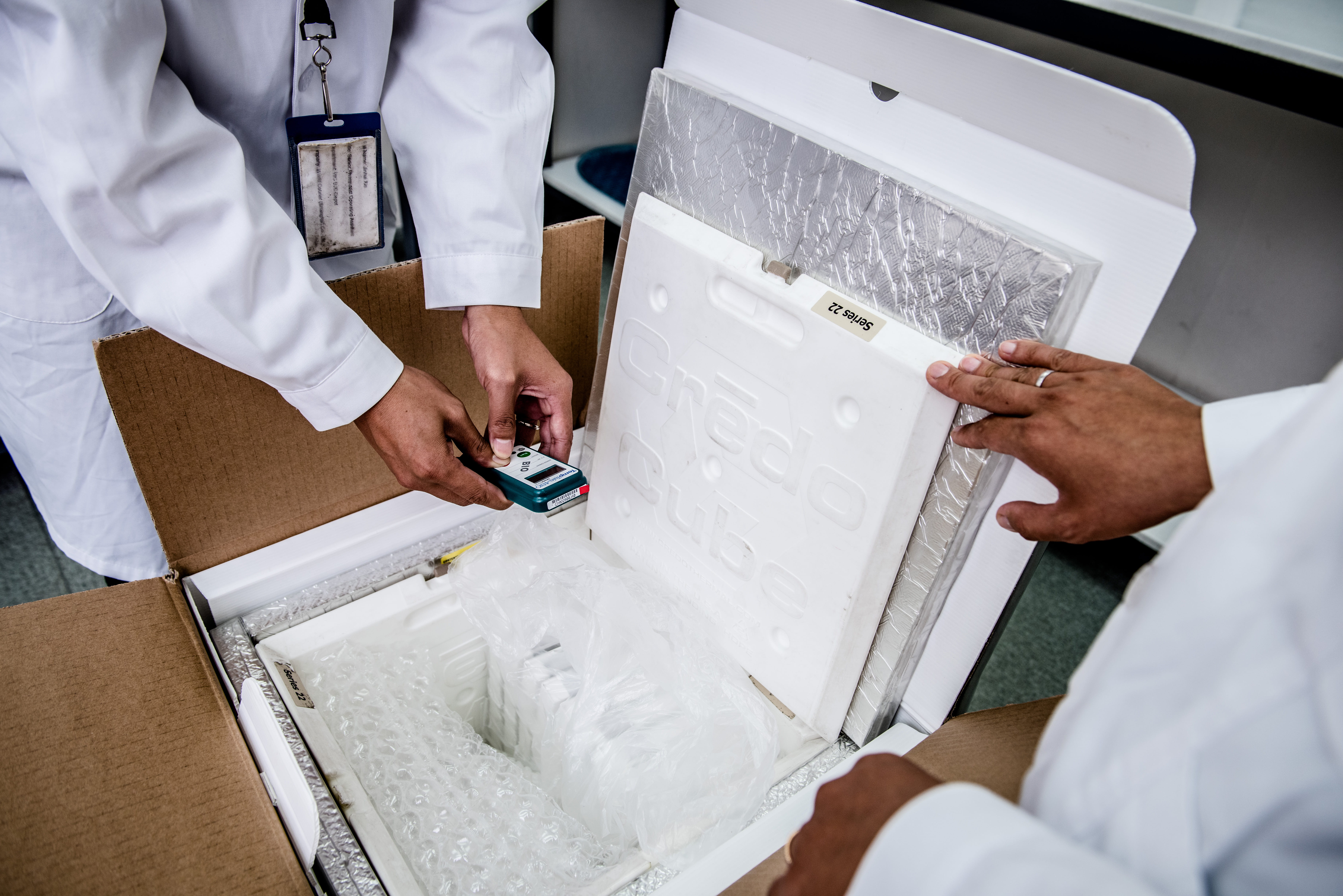 Two people in lab coats take a reading from a digital monitor inside an insulated package.