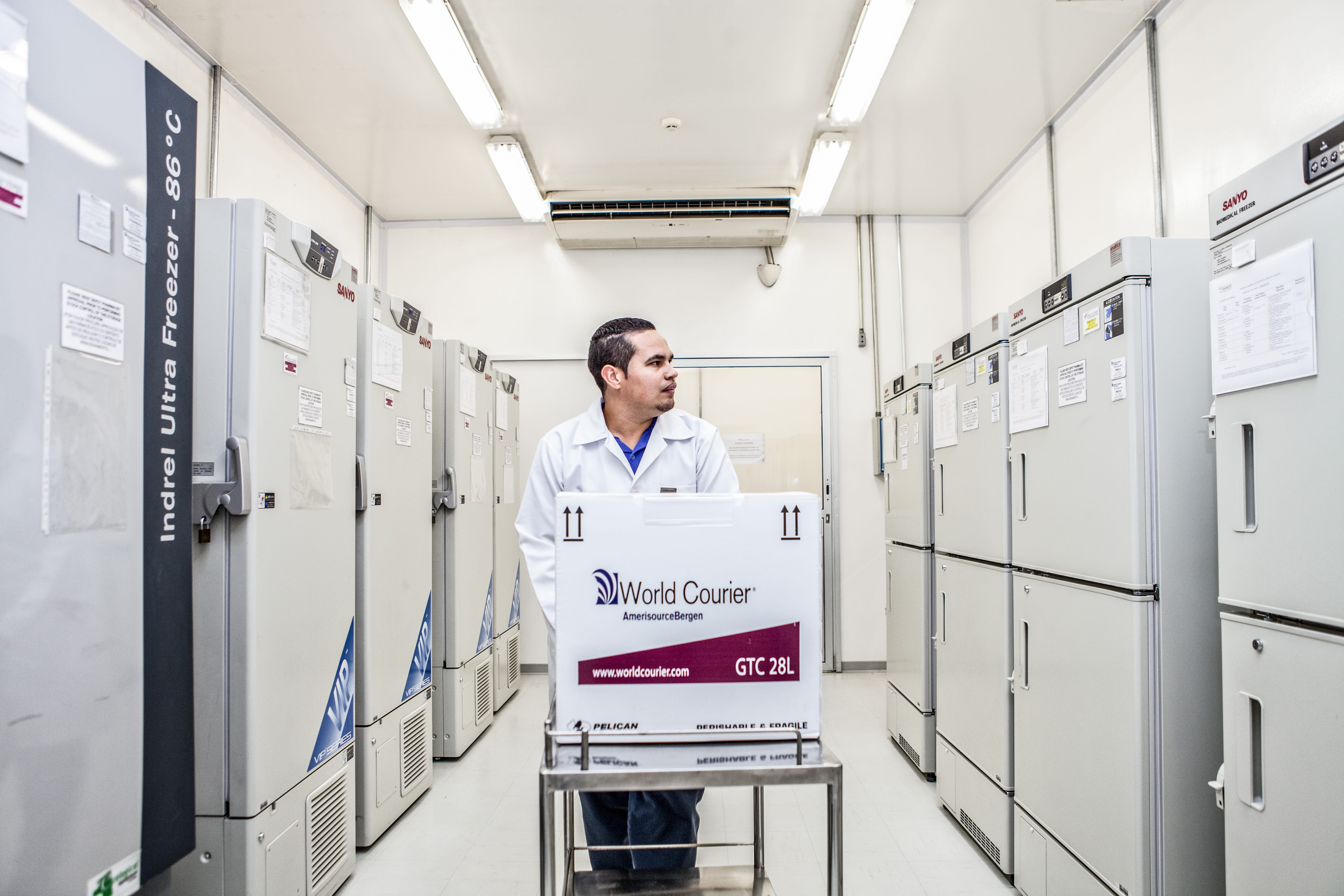 A man pushes a box on a cart down a wide aisle of storage lockers in a pharmaceutical storage depot.