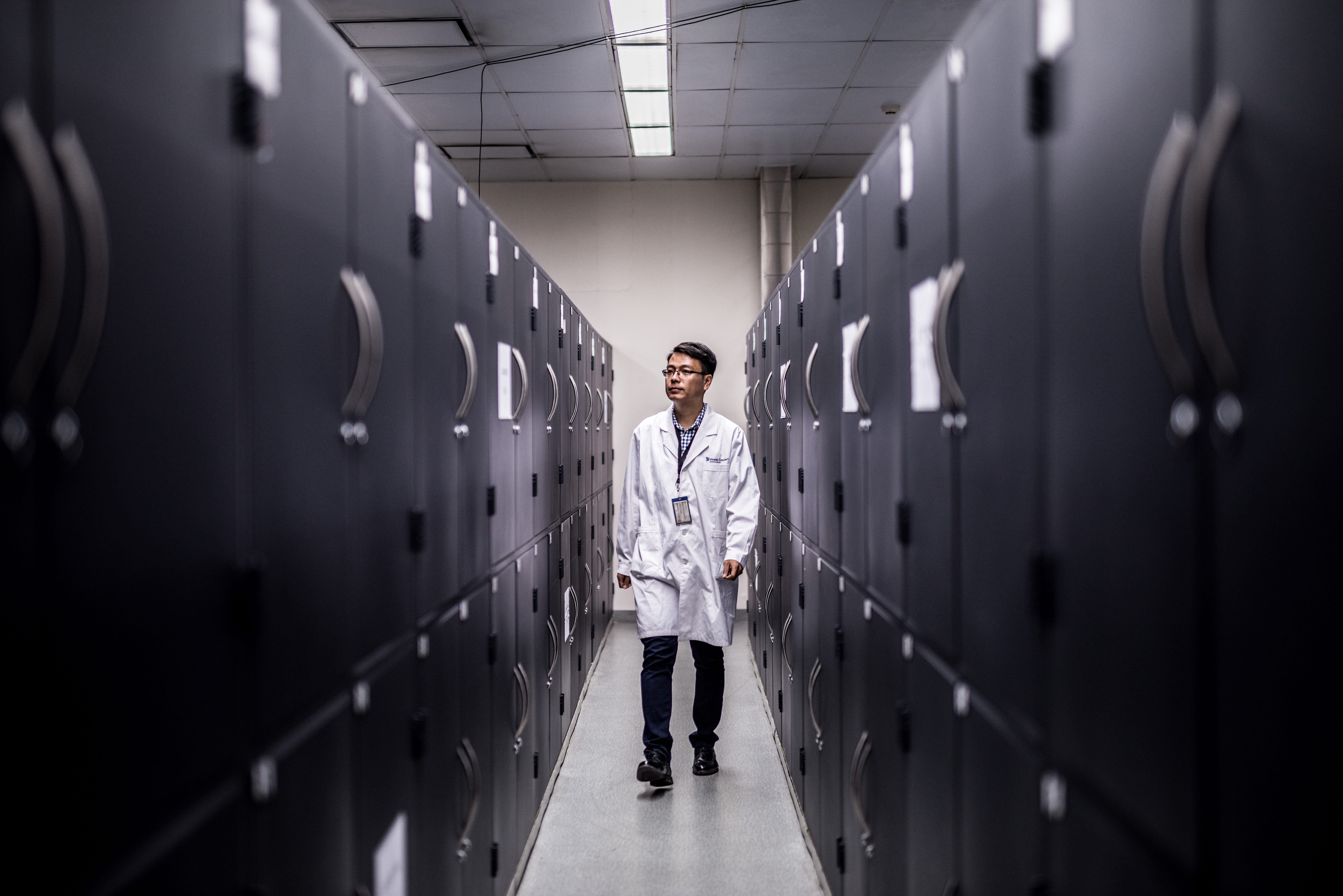 A man in a lab coat walks down a narrow aisle of lockers in a storage depot.