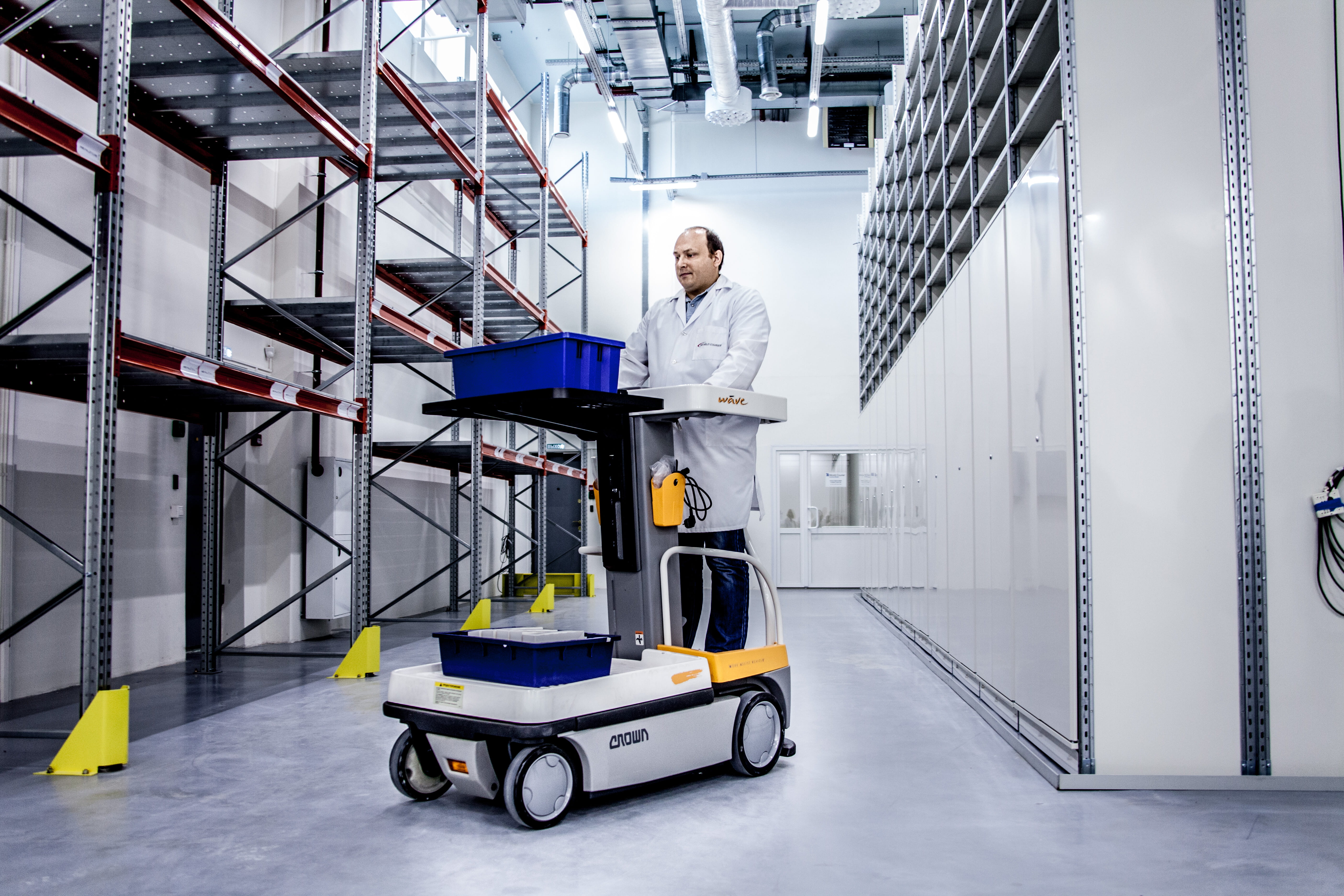 A man drives a forklift holding a small bucket down a wide aisle of shelves in a storage depot.