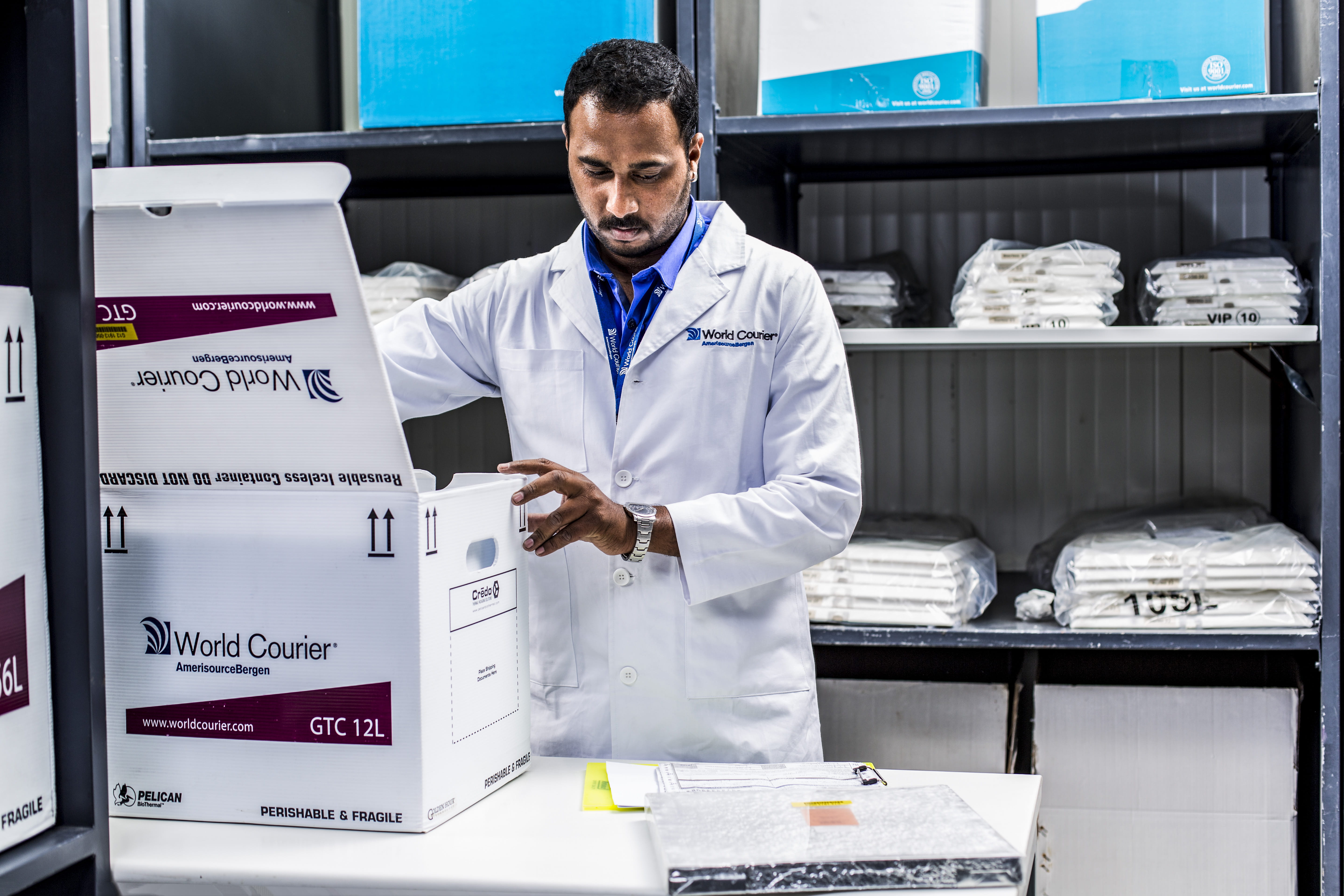 A World Courier associate in a lab coat packs a box for shipment surrounded by shelves.