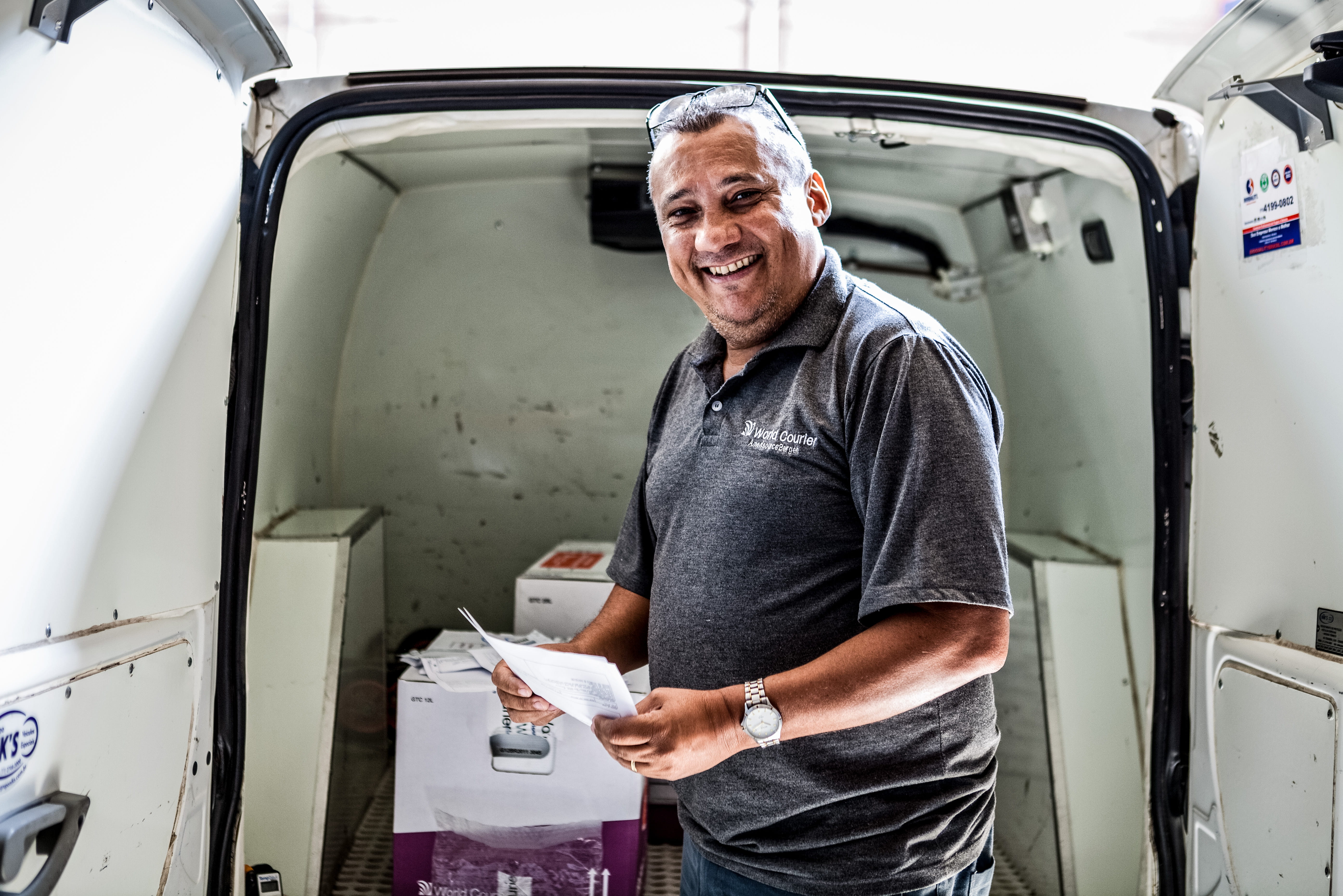 A smiling World Courier driver stands in front of his van, which is open and shows packages inside.